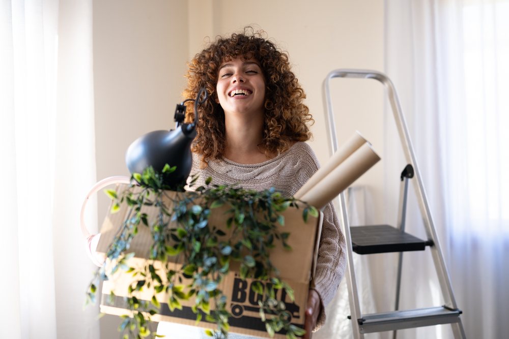 Woman smiling while carrying a cardboard box with household items and an indoor plant inside a home, moving, Spain.