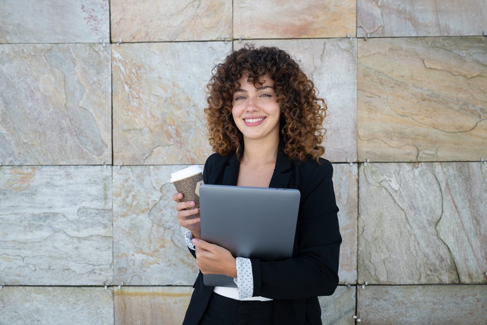 Businesswoman holding a laptop and disposable coffee cup standing in front of a stone tile wall, S, traveling executive, Spain, Antiago De Compostela.