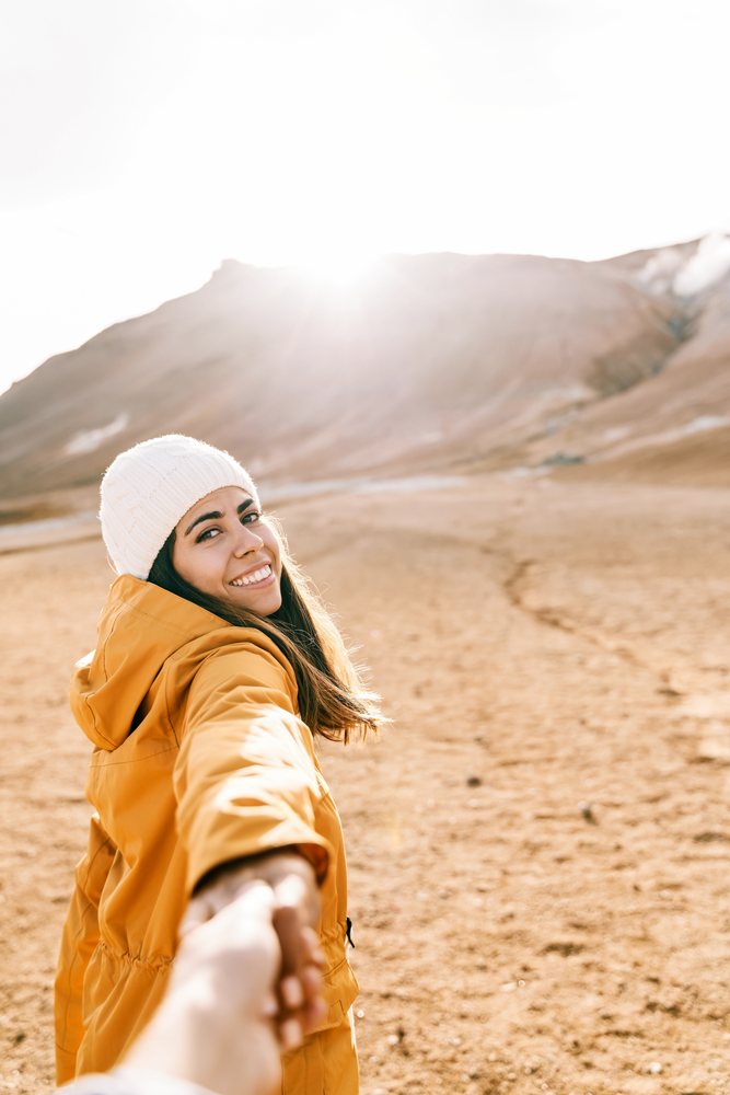 A smiling woman wearing a yellow jacket and white knit hat holds someone's hand while hiking in a sunlit mountain landscape, Iceland.