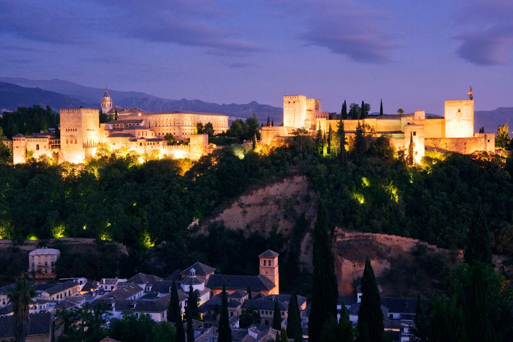 Illuminated Alhambra fortress atop a hill overlooks Granada, with mountains and cypress trees in the distance at twilight, Spain.