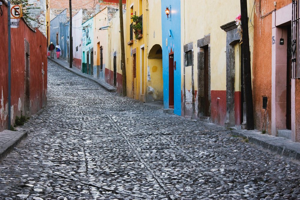 Colorful buildings line a cobblestone street with pedestrians walking by a street lamp, Guanajuato, Mexico.
