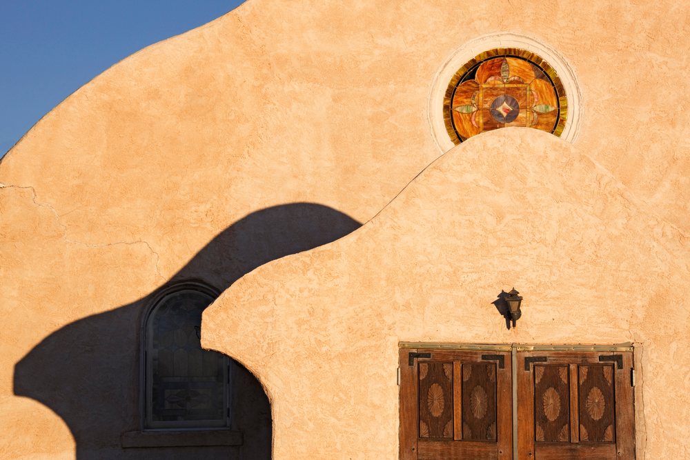 Sunlit adobe church exterior with wooden doors and stained glass windows, New Mexico, USA.