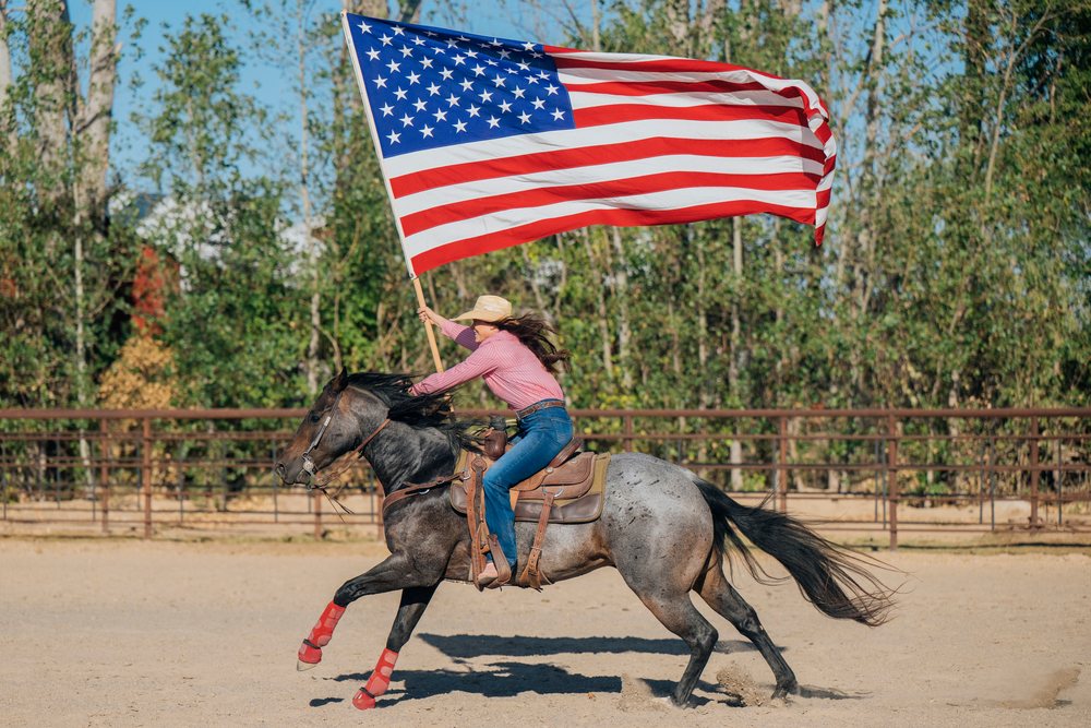 Woman rides a horse carrying a large American flag in a fenced outdoor arena, Southwest USA.