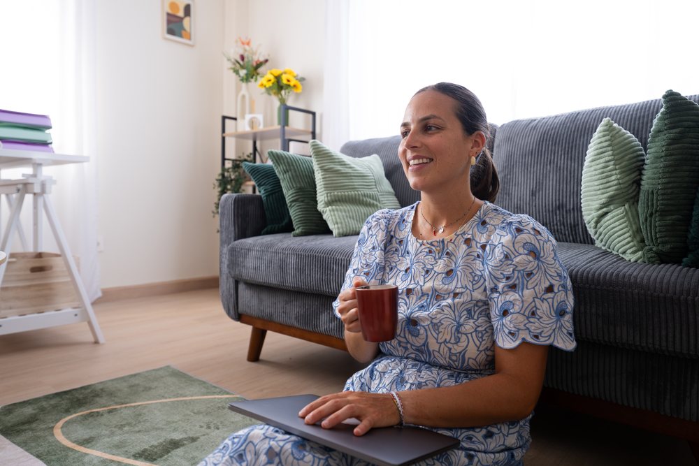 Woman smiling while holding a cup and sitting on the floor beside a laptop and gray sofa in a sunlit living room, telecommuting, Spain.