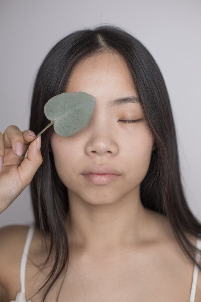 Woman with long black hair holds a leaf over one eye with a peaceful expression in a neutral studio portrait, Art, emotional.