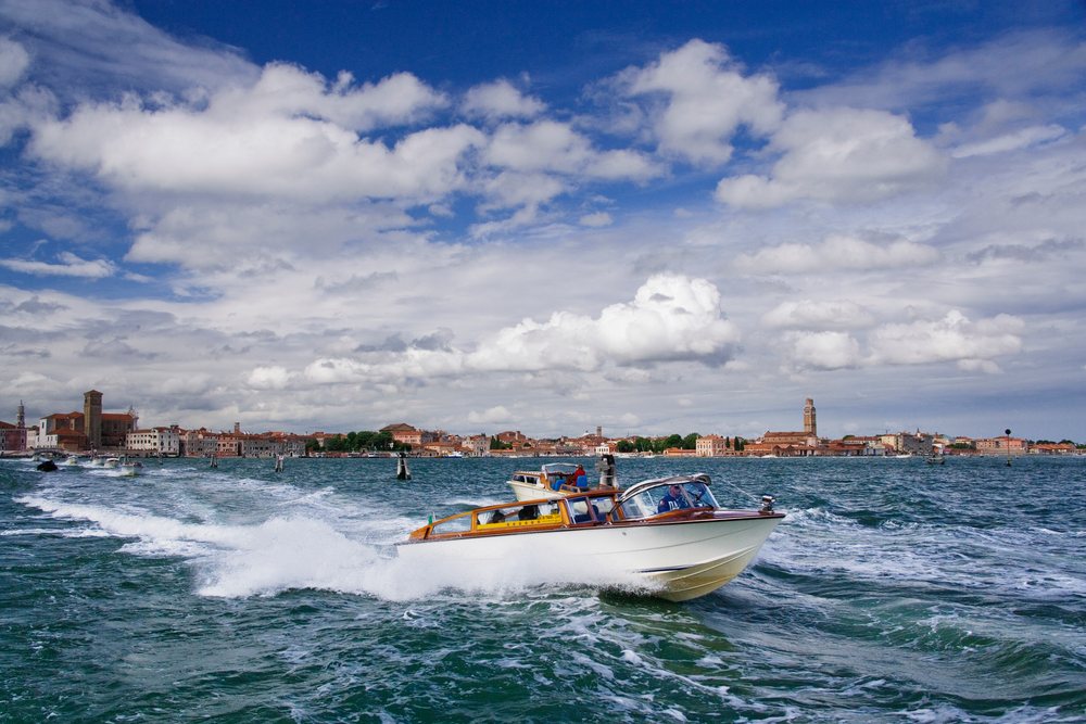Water taxi speeds across a lagoon with urban Venice buildings and a partly cloudy sky in the background, Italy.