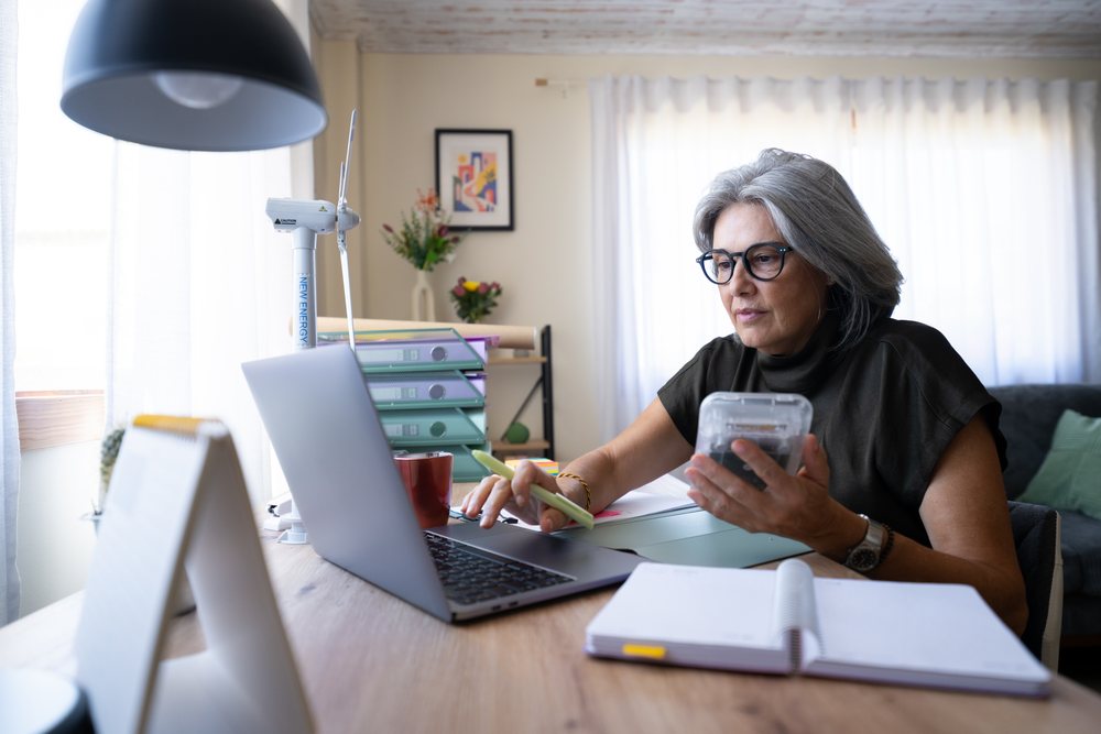 Mature woman with gray hair and glasses working at a desk using a laptop and calculator in a modern home office, Spain.