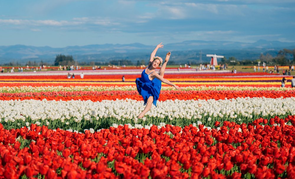Ballet dancer in blue dress wearing a flower crown poses amid vibrant tulips with a windmill and mountains visible under a cloudy blue sky, USA.