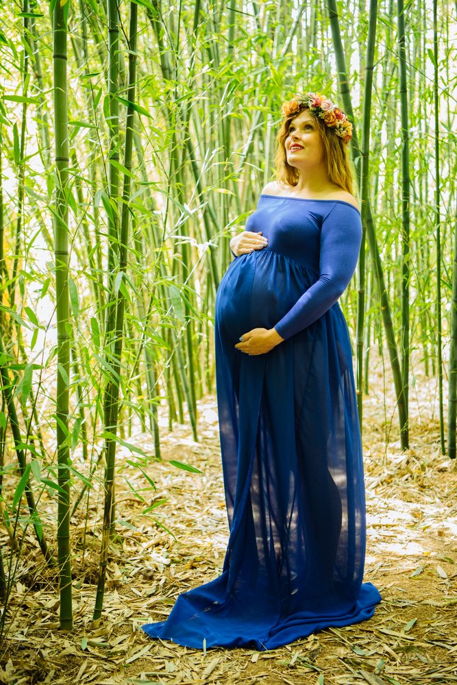Pregnant woman wearing a flower crown and flowing blue dress stands outdoors among bamboo stalks with hands on her belly, pregnancy.