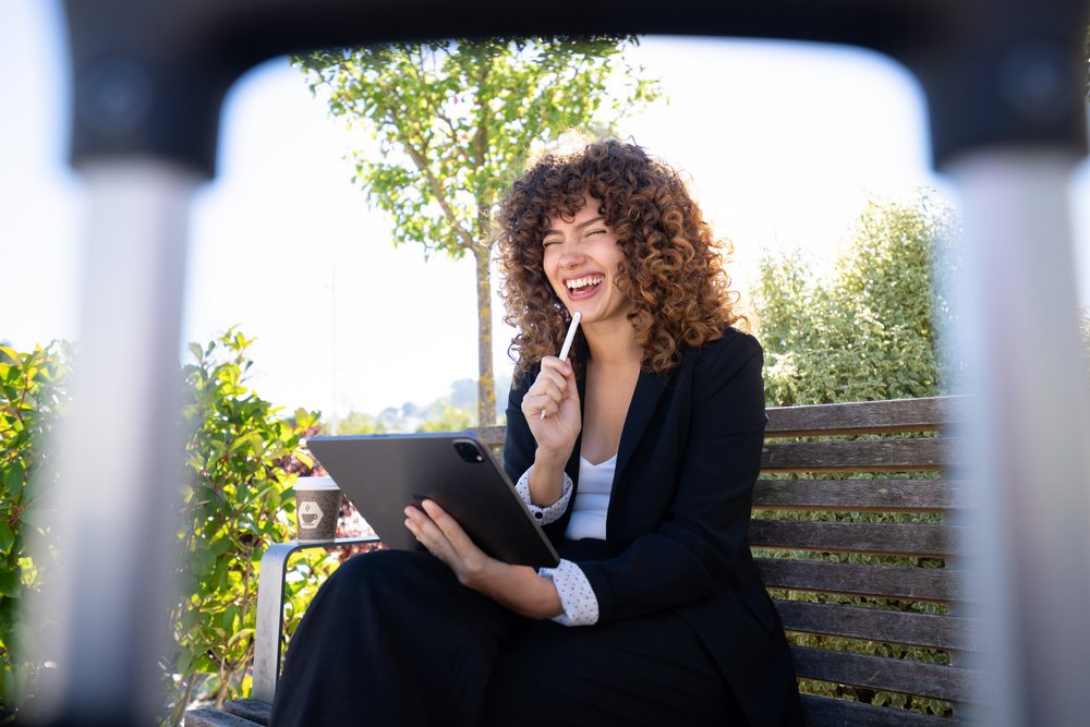 Woman sitting on a wooden bench outdoors in daylight, laughing while holding a tablet and pen near greenery with a coffee cup on the bench armrest, Spain.