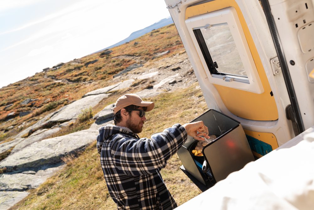 Man cooking food on a portable camping stove at the rear of a camper van in a rocky outdoor landscape, Norway.