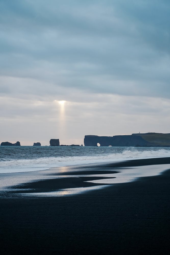 A sunbeam shines through clouds over black sand beach and ocean rocks