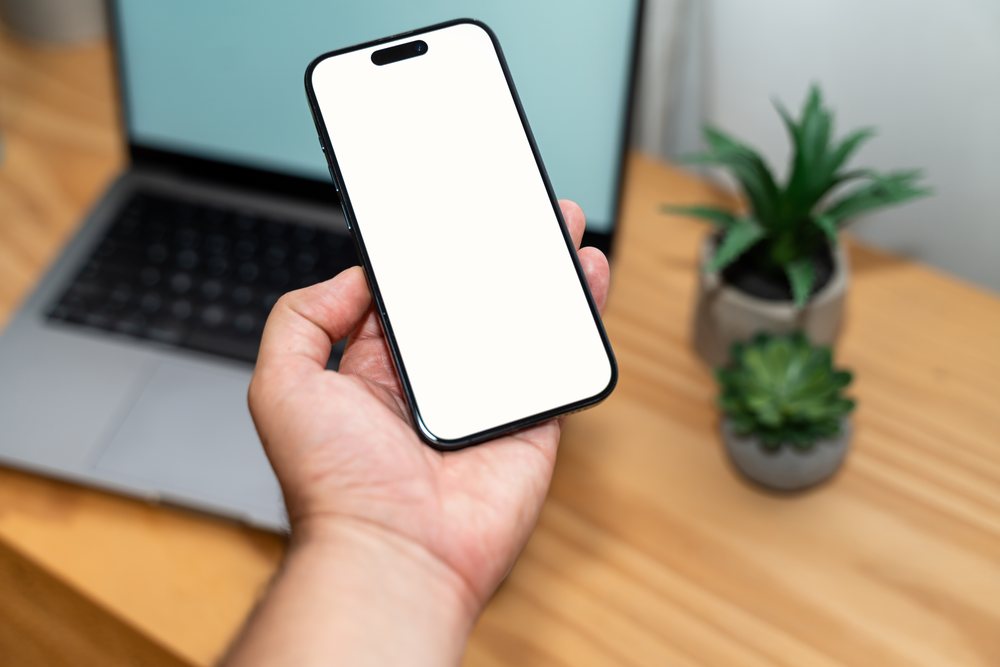 Smartphone with blank white screen held in left hand on a wooden desk near laptop and two potted succulent plants in an indoor workspace, mock up, Spain.