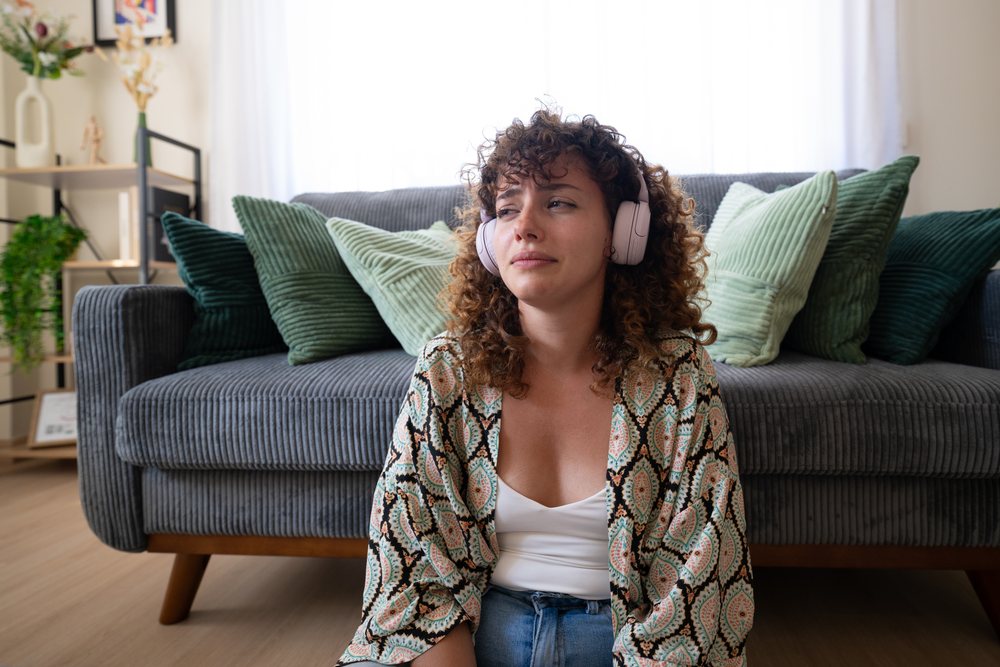 Woman with curly hair wearing headphones sitting on a wooden floor in front of a couch with cushions in a living room, home, girl, Spain.