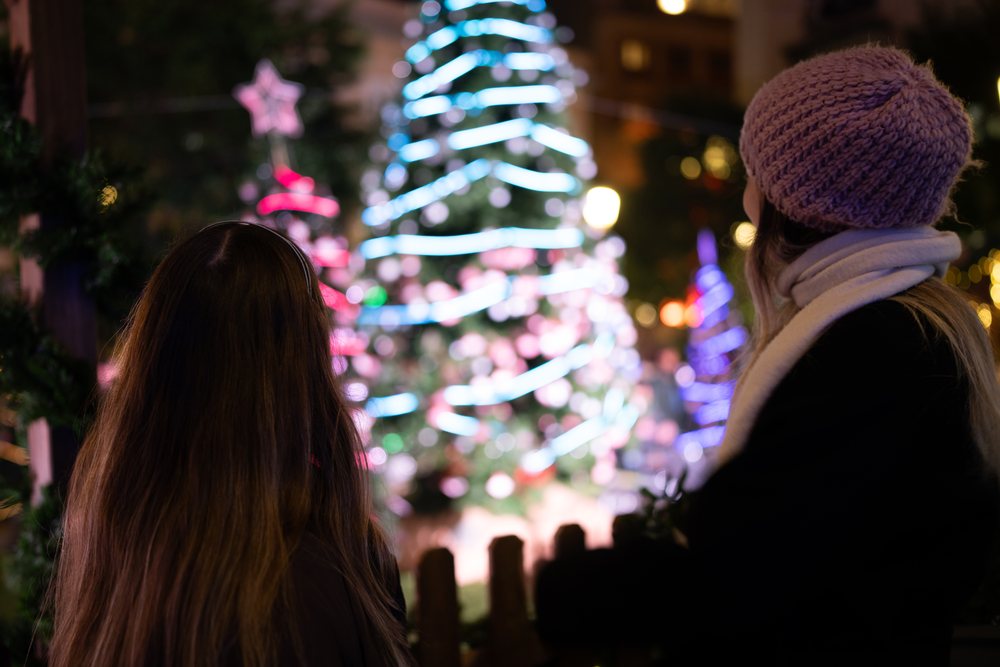 Two women wearing winter clothing admire a decorated and illuminated Christmas tree at a nighttime Christmas market, Madrid, Spain.