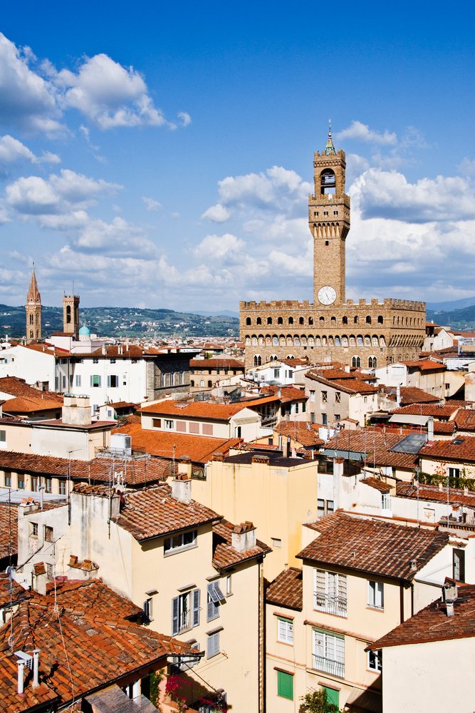 Palazzo Vecchio clock tower and red tile rooftops under a partly cloudy blue sky, Florence, Italy.