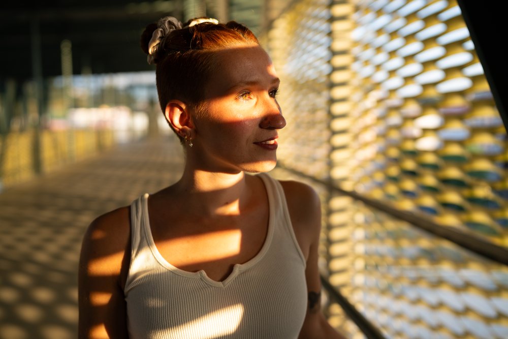 A young woman stands in a modern urban walkway with striking sunlight and shadows creating a light pattern on her face and body, Santiago De Compostela, Spain.