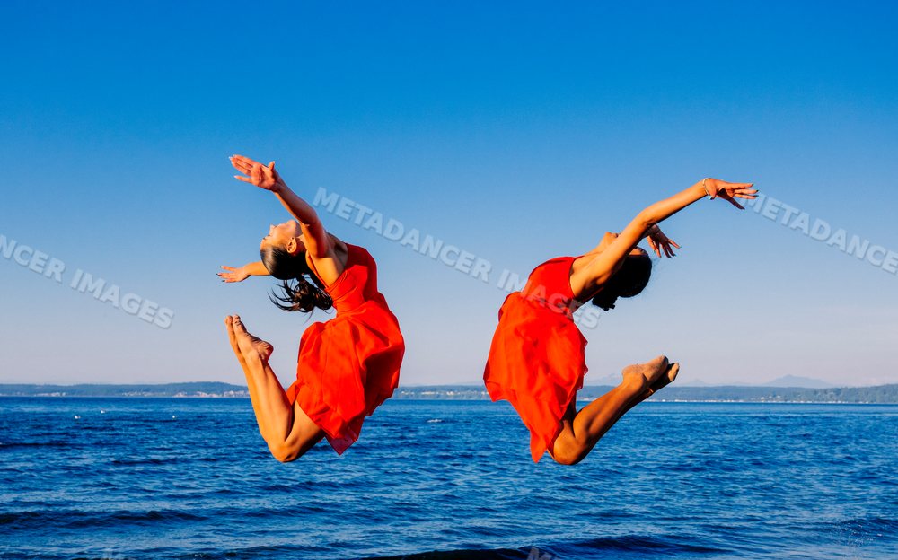 Two women in red dresses jump in acrobatic poses by the ocean shore