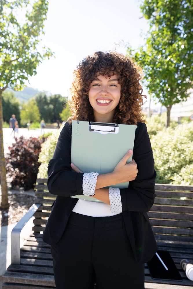 Smiling woman with curly hair sits on a wooden park bench outdoors holding a green clipboard on a sunny day, Spain.