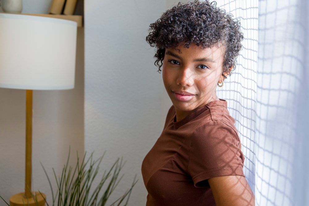 Young woman with curly hair smiling near a window inside a home.