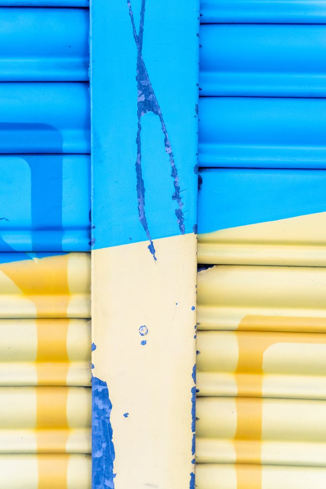 Close-up of a weathered metal shutter with peeling blue and yellow paint, showing surface texture and wear in an urban setting, abstract texture, background, wallpaper, Spain.