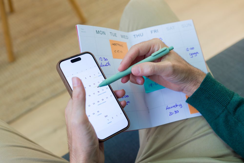 Person writing an appointment in a paper calendar planner while holding a smartphone at home, Young adult.
