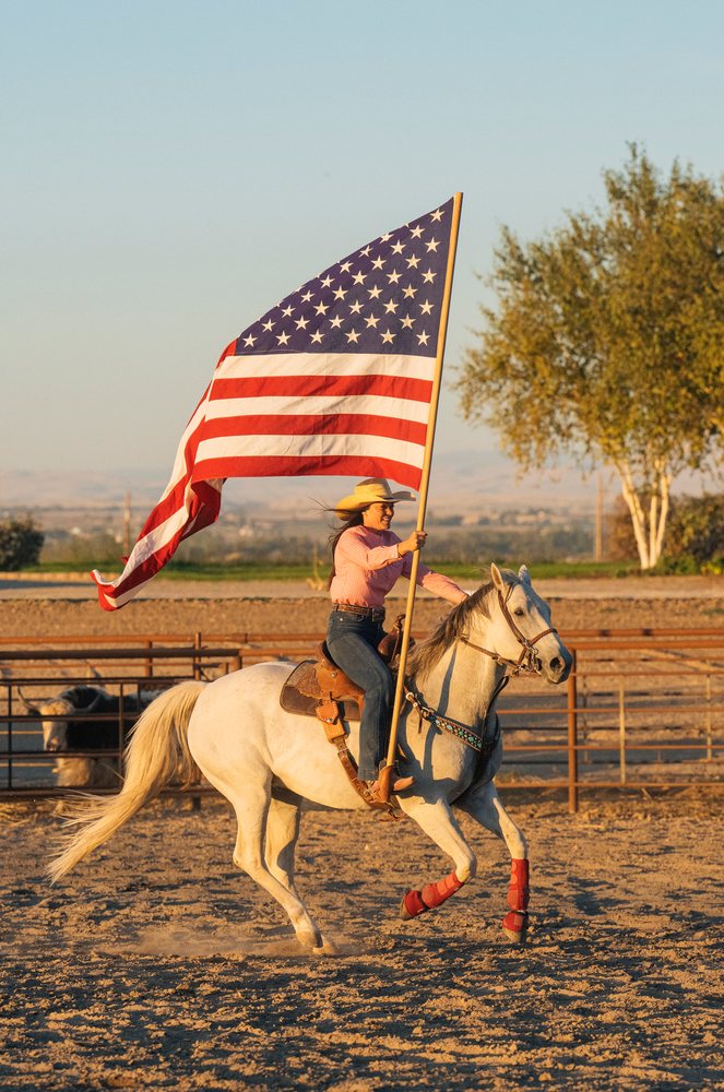Woman wearing a cowboy hat rides a white horse in a fenced outdoor ranch arena holding a large American flag, Southwest USA.