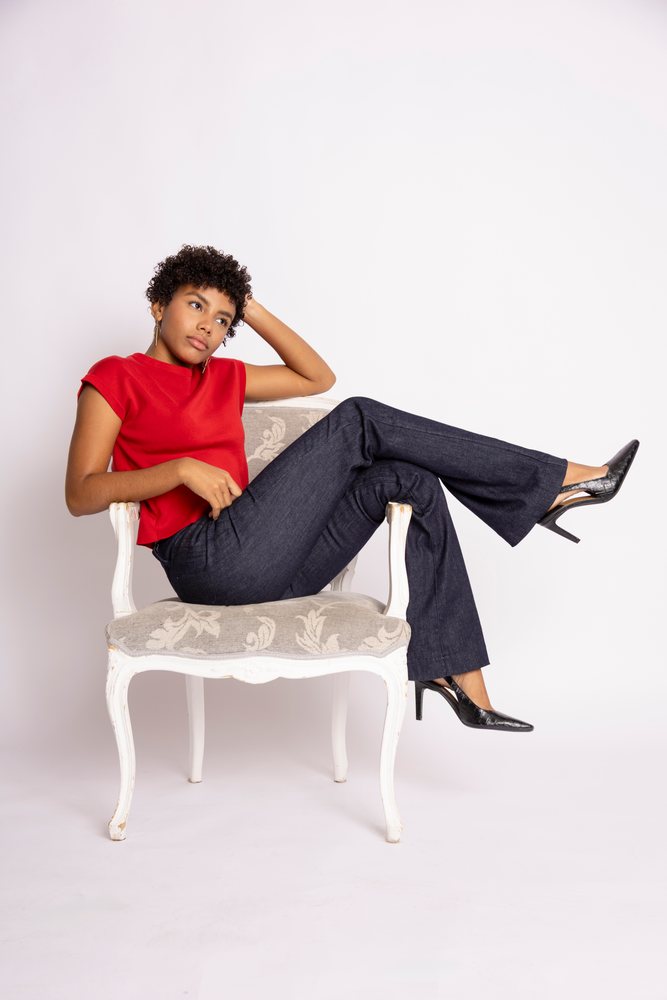 Young woman with curly hair wearing a red top and black pants sitting relaxed on an elegant white armchair against a white studio background, fashion, clothes, young culture.