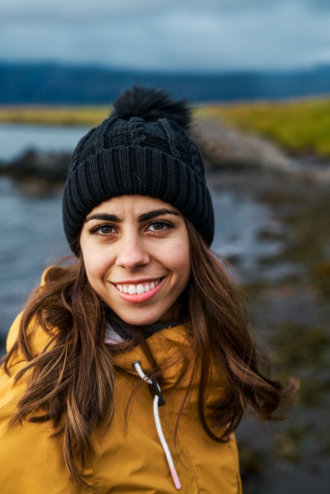 A white woman smiles outdoors in cold weather near a rocky shore