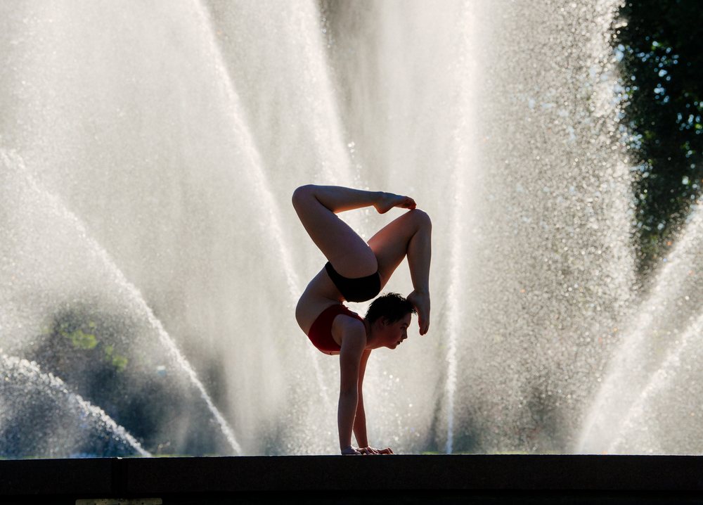Young adult woman performs a flexible handstand on a stone ledge with a large water fountain spraying behind her outdoors, USA.