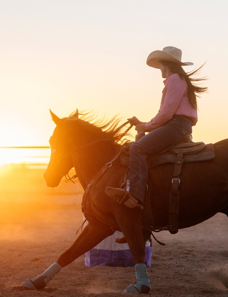 Cowgirl wearing a western hat rides a brown horse with hoof protection in an outdoor arena at sunset with dust swirling around, Southwest USA.