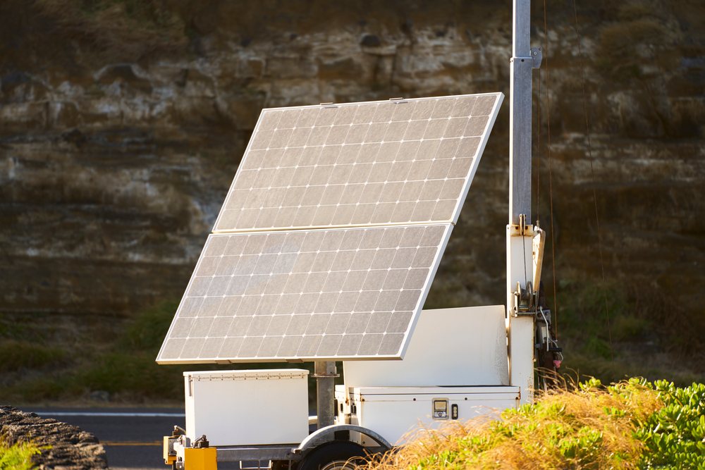 Two large solar panels mounted vertically on a white trailer base outdoors with sunlight and rock face background, Oahu, Hawaii, USA.