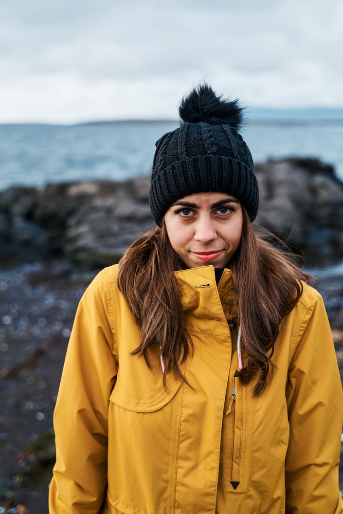 White Woman in yellow jacket and black hat on rocky beach in a beach