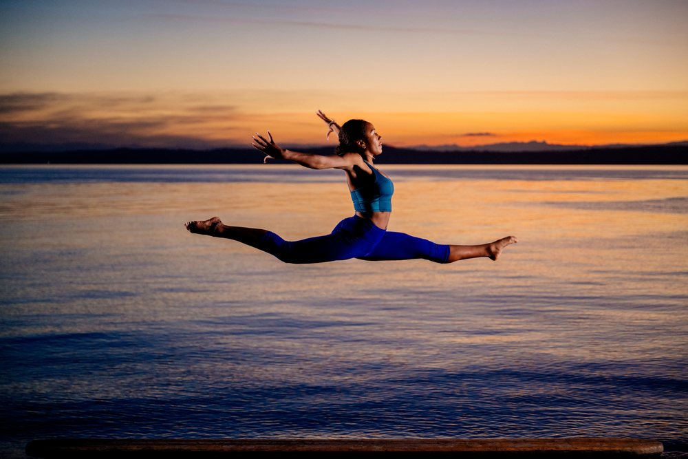 Woman performing an acrobatic split jump exercise outdoors at sunset by the water, yoga, acrobatics, USA.