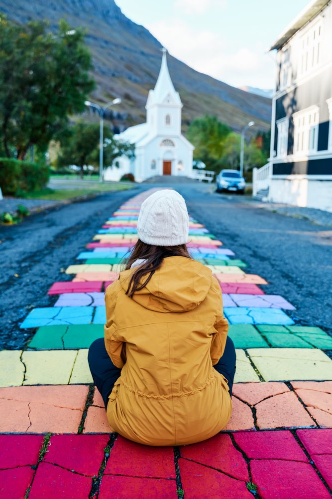 A woman dressed in a yellow jacket and white knit hat sits on a colorful paved path that leads to a small church in a scenic village, Iceland.