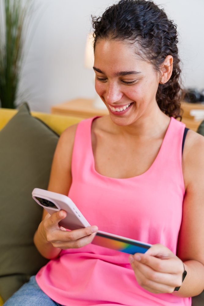 A smiling woman uses her smartphone to make an online purchase with a payment card at home, Spain.