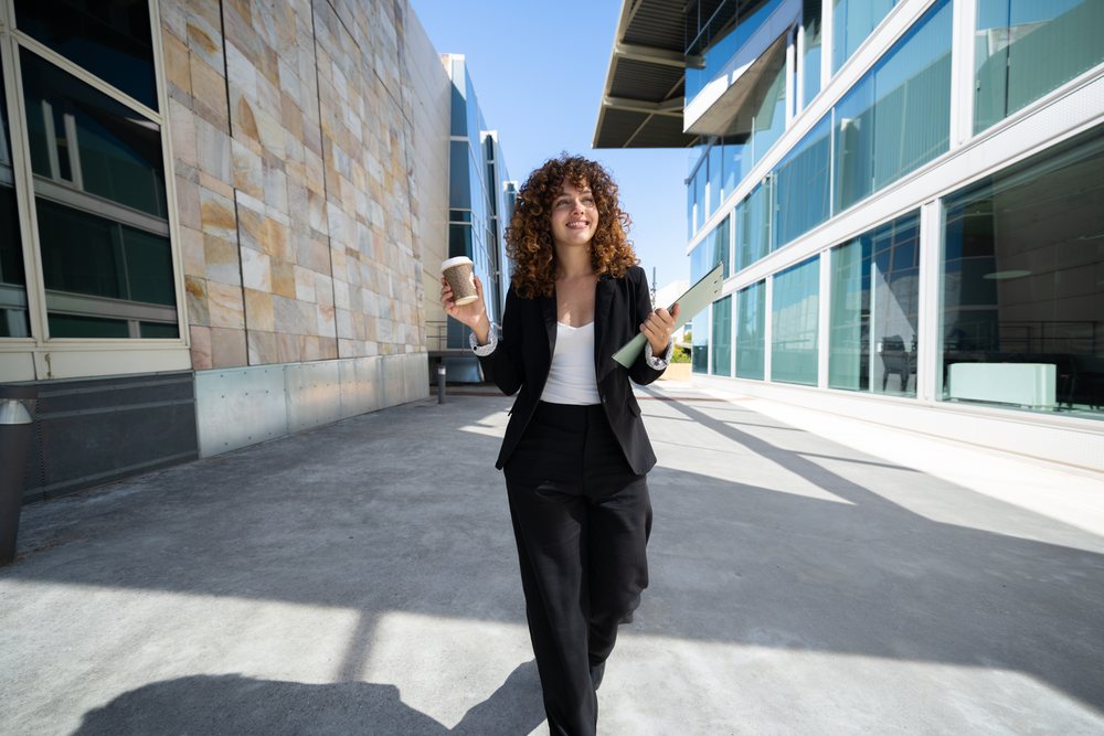 Businesswoman walking outdoors with coffee and clipboard between modern office buildings, traveling executive, Spain.