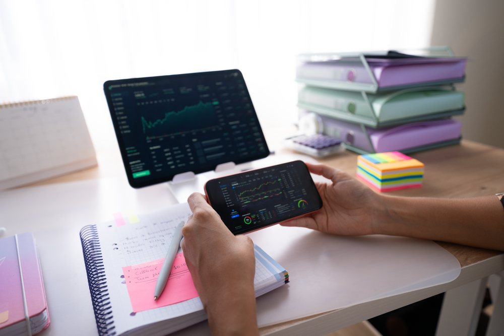 Tablet and smartphone displaying financial graphs on a desk with notebook, pen, sticky notes, and binders in a home office, Spain.