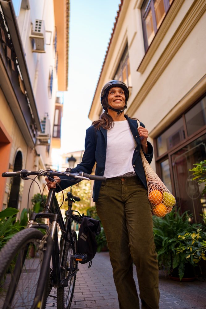 Woman wearing helmet walking and holding a bicycle on a street in Málaga carrying a net bag with oranges and a lemon.