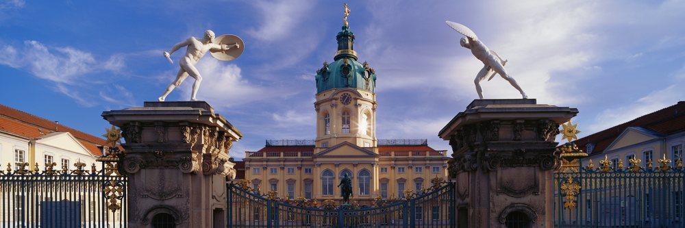 Charlottenburg Palace entrance gate with statues, clock tower, and ornate fence under a clear blue sky, Charlottenburg Palace Entrance Berlin, Germany.