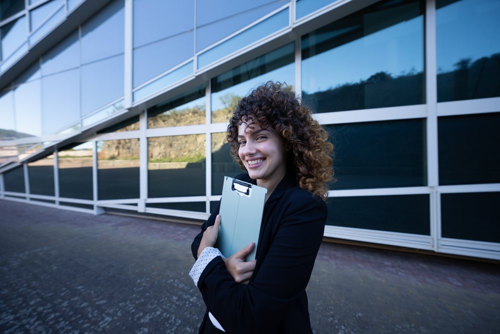 Smiling businesswoman with curly hair holds a clipboard outside a modern glass building, business trip, Spain, Santiago De Compostela.