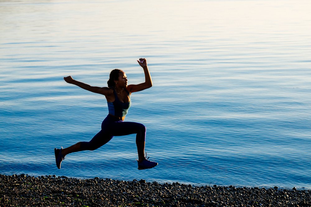 Woman jumping in an athletic pose on a pebble beach by a calm lake during outdoor exercise, yoga, acrobatics, USA.