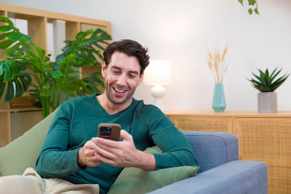 Man smiling while sitting on a sofa and using a smartphone in a modern living room with indoor plants, Technology, young adult, home.