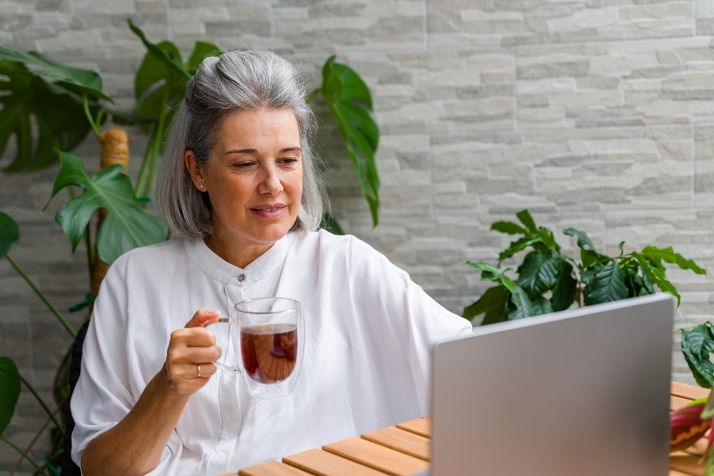 Mature woman with gray hair drinking tea and looking at a laptop in a modern workspace with indoor plants, Spain.