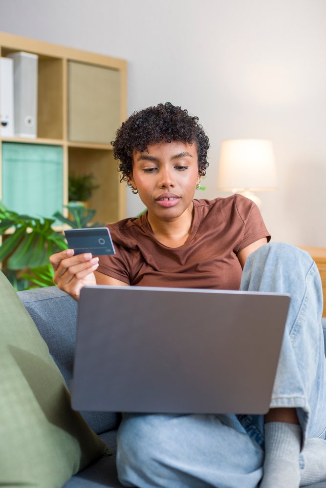Young woman sitting on couch at home using laptop and holding credit card with modern interior elements visible.