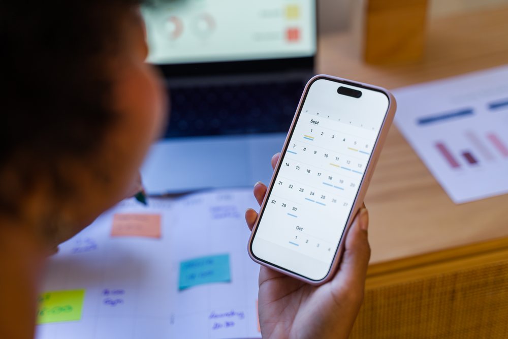 Person holding smartphone displaying calendar app while sitting at desk with planner, sticky notes, laptop, and charts in Spain office.