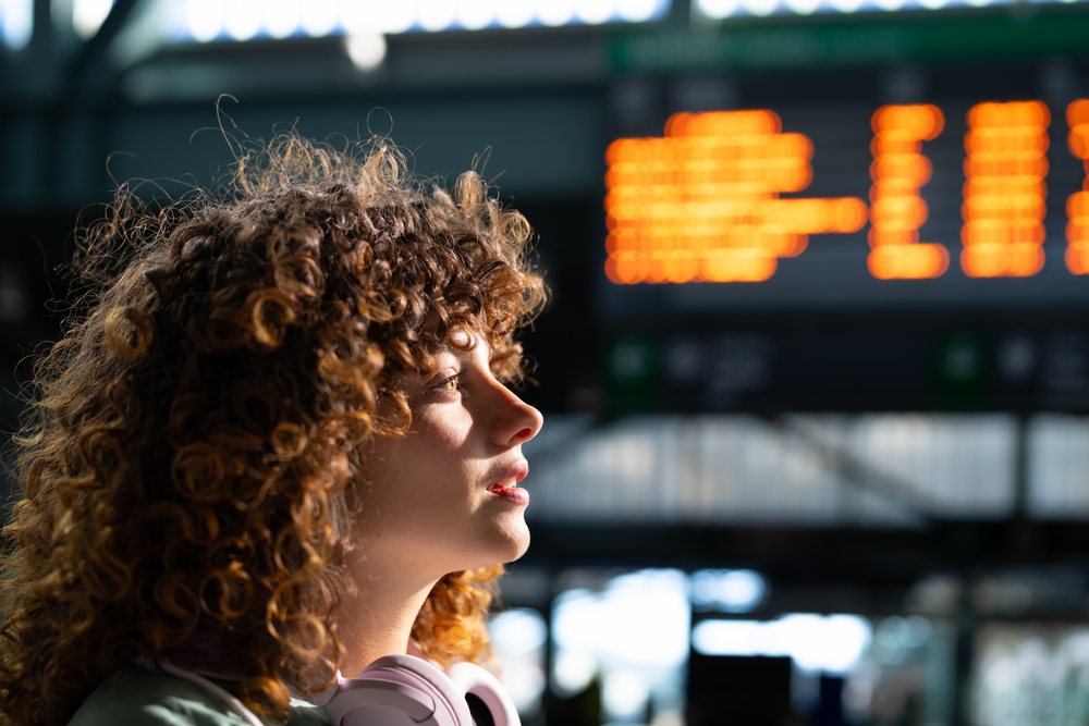 Woman with curly hair wearing headphones waits indoors at a train station with an illuminated departure board in the background, Spain.