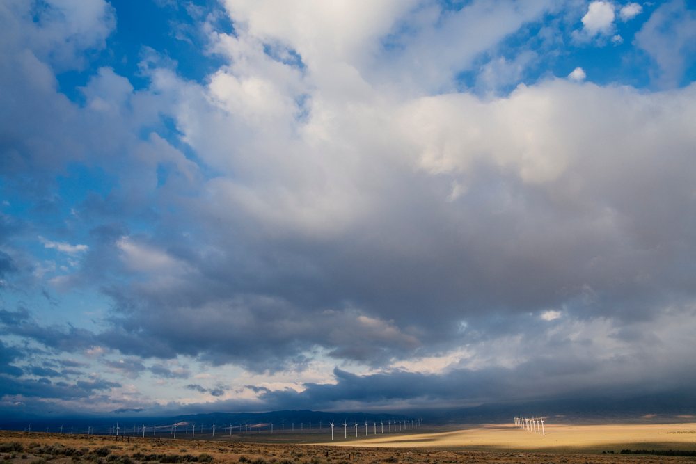 Wind turbines stand across a flat, dry Nevada landscape beneath a cloudy sky with distant mountains, USA.