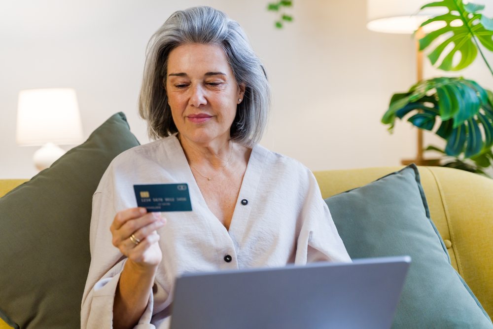 Middle-aged woman with gray hair holds a credit card and uses a laptop while seated on a sofa in a living room, Spain.