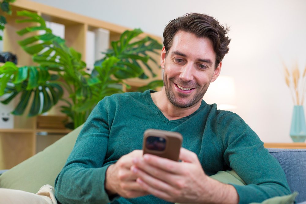 Man sitting on a couch using a smartphone in a living room with plants, a lamp, and a shelf with folders, Technology, young adult, home.